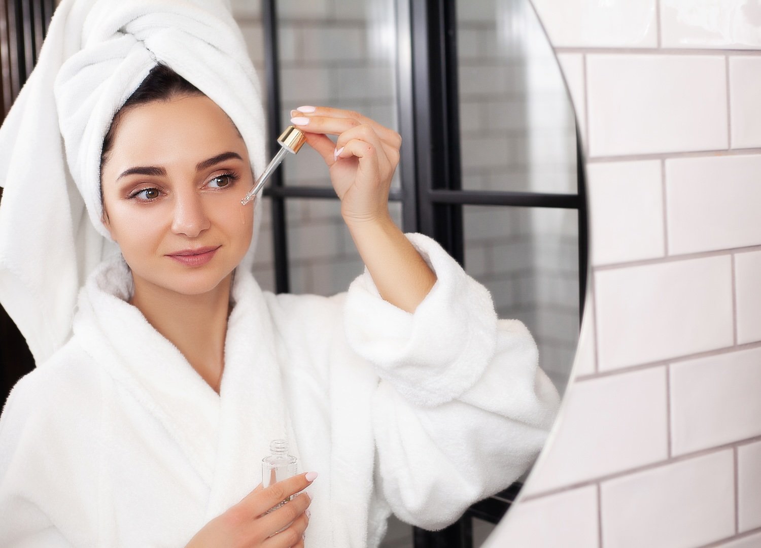 A woman wearing a bathrobe uses a face serum and examines her reflection in the mirror